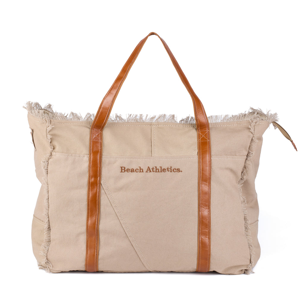 Bonnie: a beige washed canvas beach bag with frayed raw edges, tan leather handles and trim, and an embroidered Beach Athletics logo, front view with the handles raised