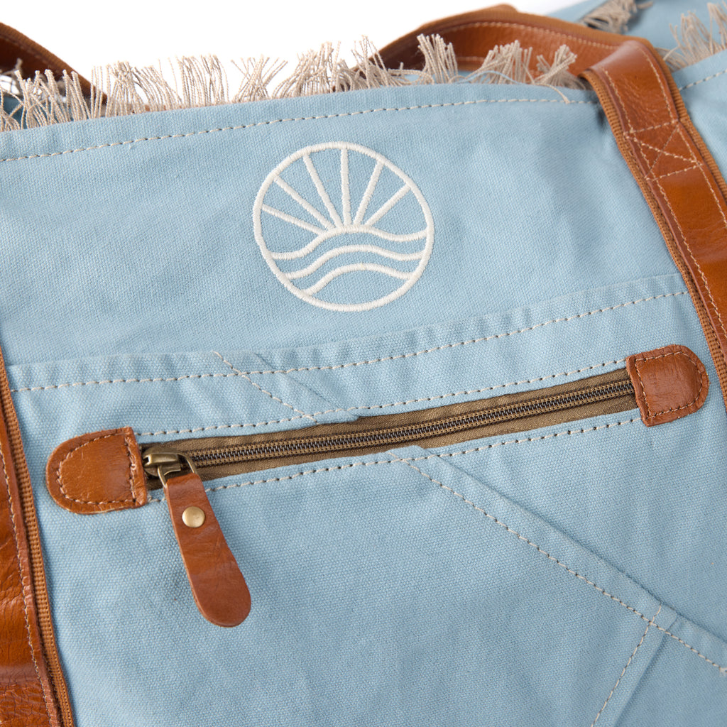 Bonnie: a blue washed canvas beach bag with frayed raw edges, tan leather handles and trim, and an embroidered Beach Athletics logo, back view