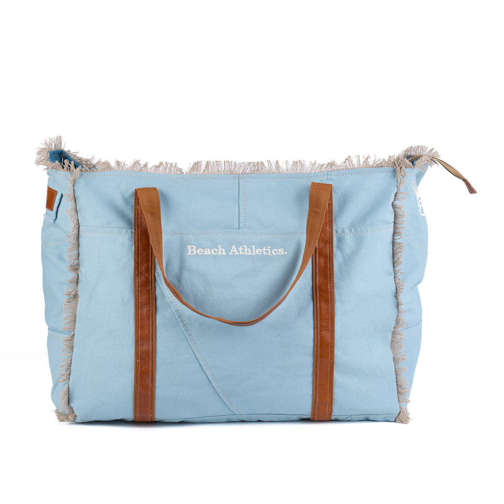 Bonnie: a blue washed canvas beach bag with frayed raw edges, tan leather handles and trim, and an embroidered Beach Athletics logo, front view, second angle