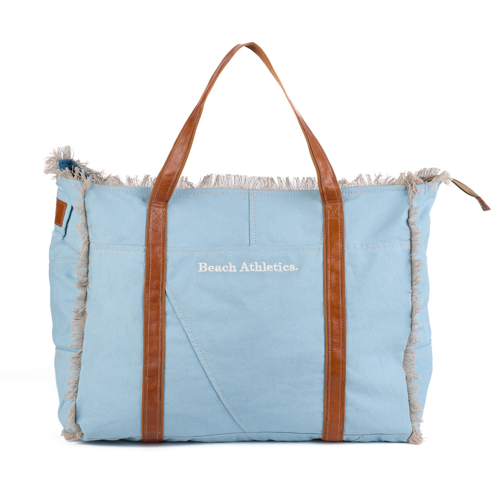 Bonnie: a blue washed canvas beach bag with frayed raw edges, tan leather handles and trim, and an embroidered Beach Athletics logo, front view with the handles raised