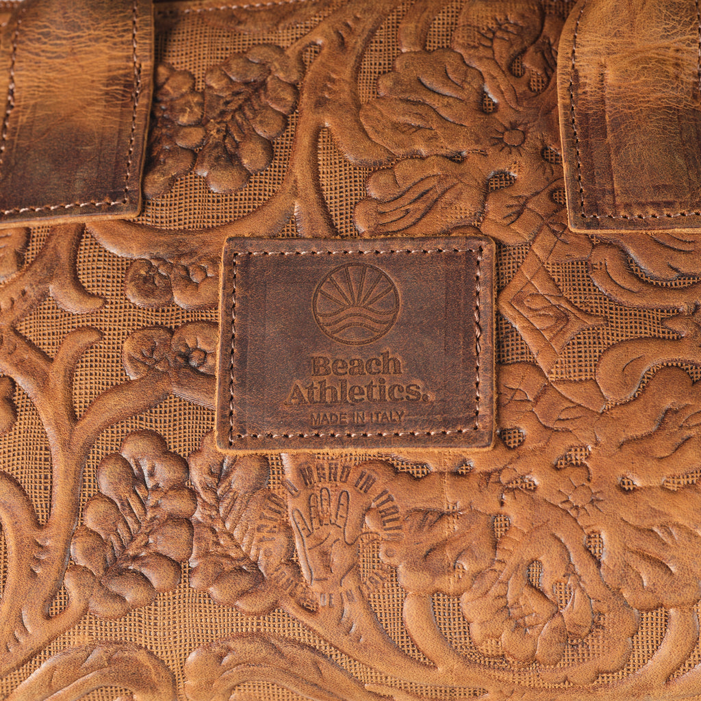 Close-up of a brown leather bag with floral tooling and an embossed central patch reading Beach Athletics Made in Haw.
