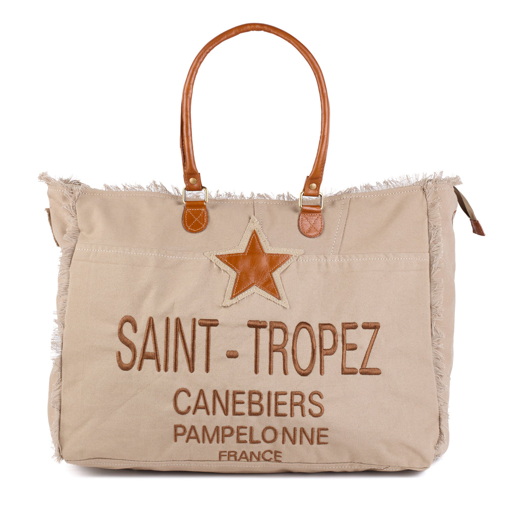 Casey: a beige washed canvas beach bag with frayed raw edges, a tan leather star patch and embroidered Saint-Tropez Canebiers Pampelonne France lettering, front view with the handles raised