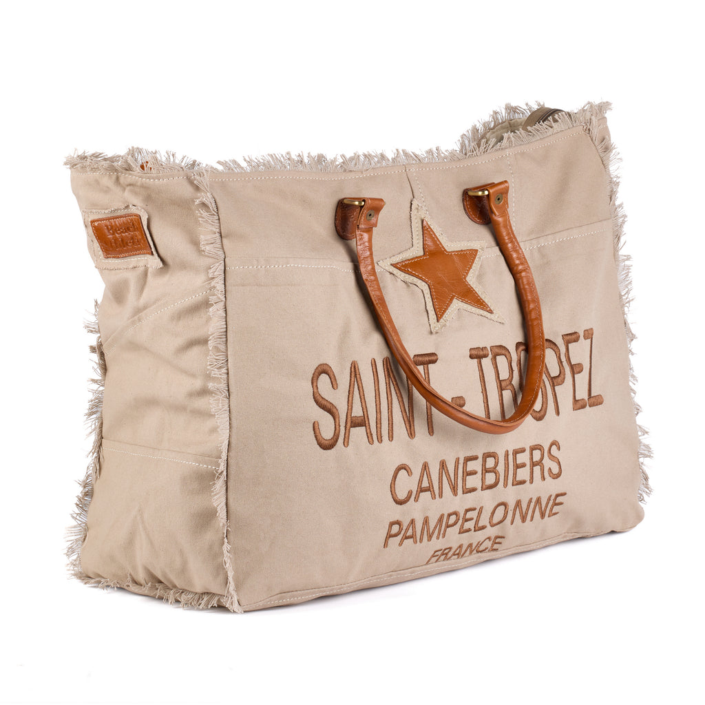 Casey: a beige washed canvas beach bag with frayed raw edges, a tan leather star patch and embroidered Saint-Tropez Canebiers Pampelonne France lettering, shown at an angle