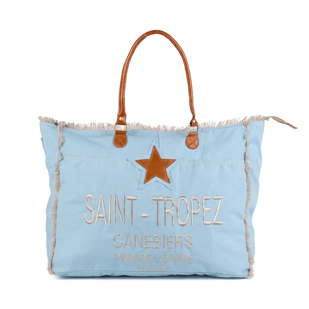Casey: a blue washed canvas beach bag with frayed raw edges, a tan leather star patch and embroidered Saint-Tropez Canebiers Pampelonne France lettering, front view with the handles raised