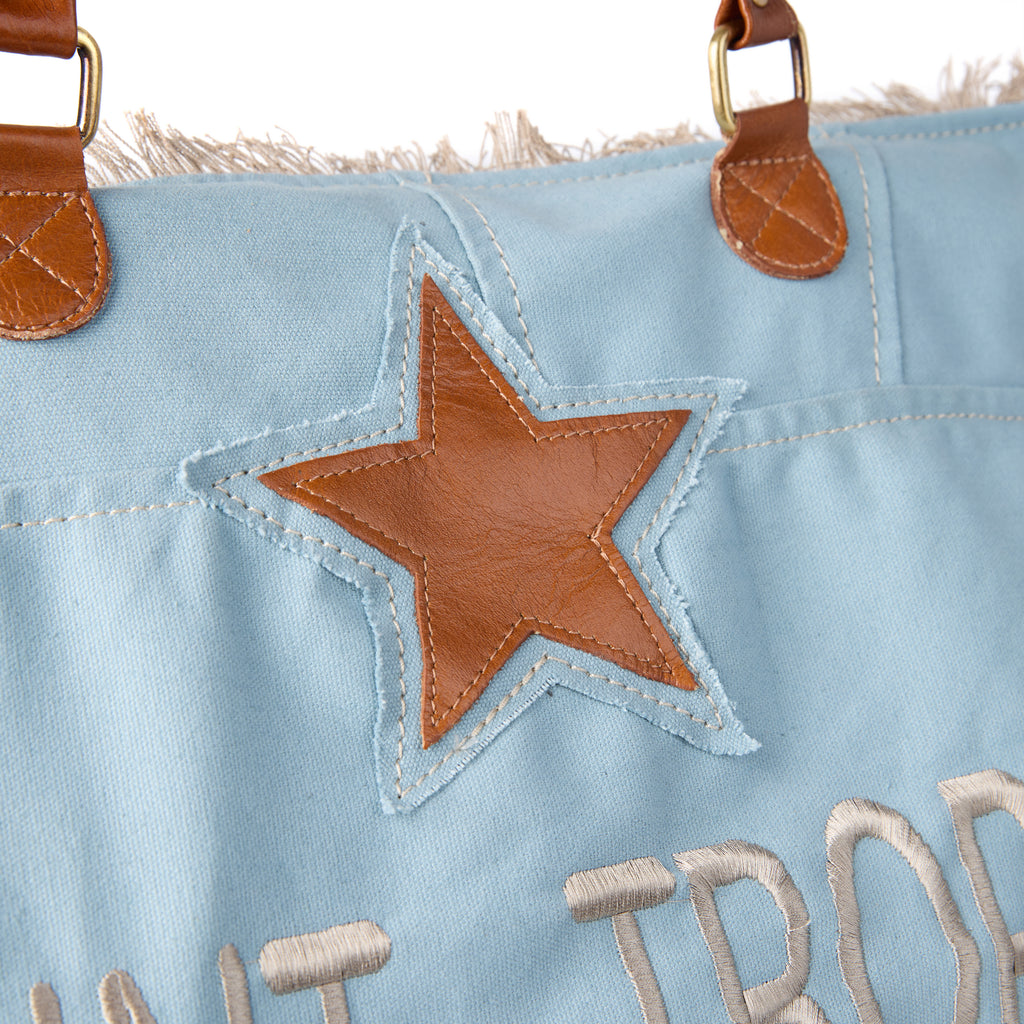 Casey: a blue washed canvas beach bag with frayed raw edges, a tan leather star patch and embroidered Saint-Tropez Canebiers Pampelonne France lettering, shown at an angle