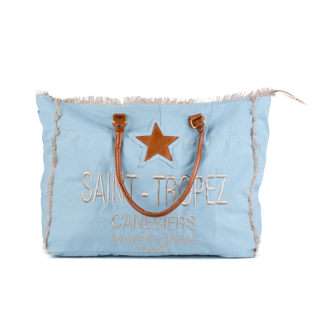 Casey: a blue washed canvas beach bag with frayed raw edges, a tan leather star patch and embroidered Saint-Tropez Canebiers Pampelonne France lettering, front view