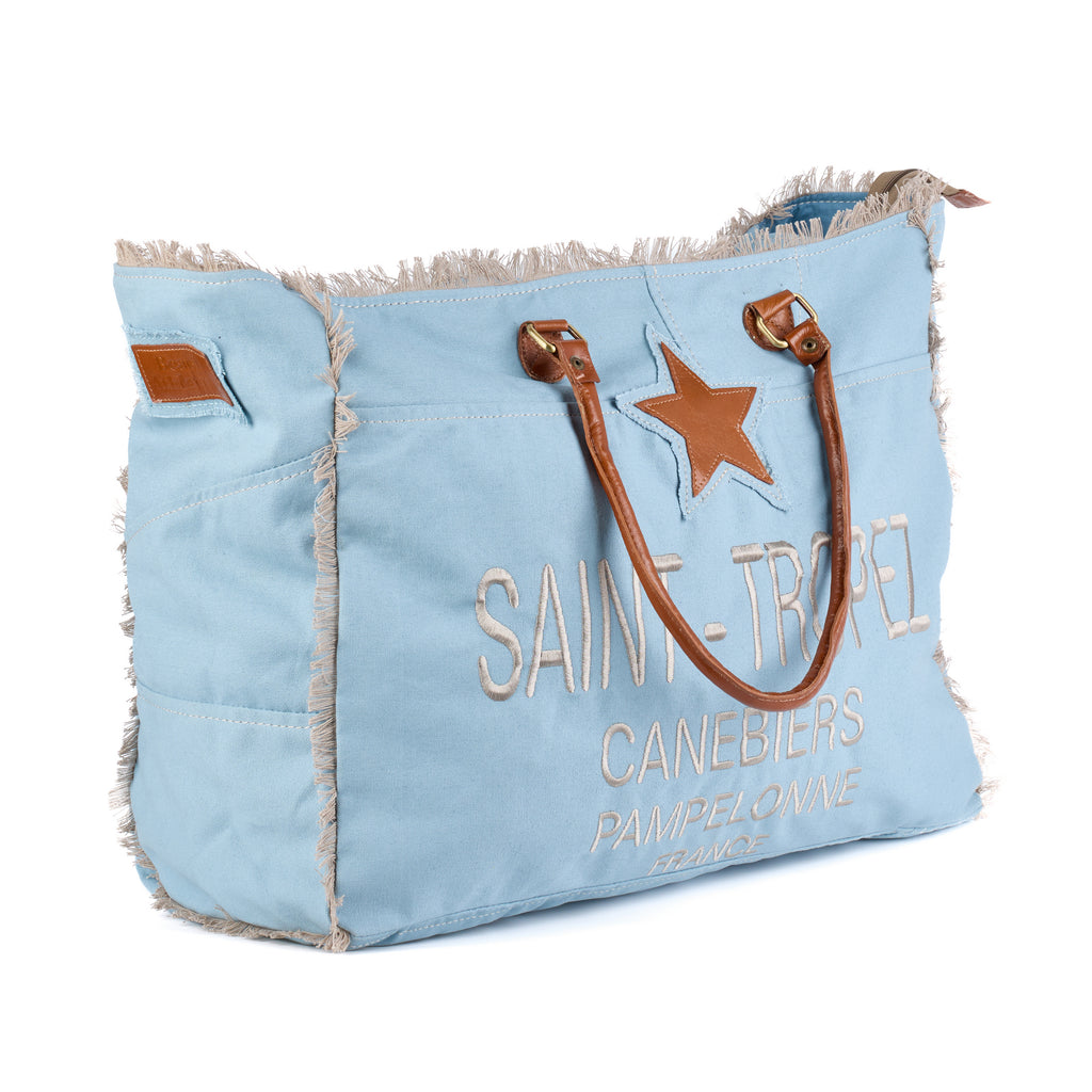 Casey: a blue washed canvas beach bag with frayed raw edges, a tan leather star patch and embroidered Saint-Tropez Canebiers Pampelonne France lettering, shown at an angle