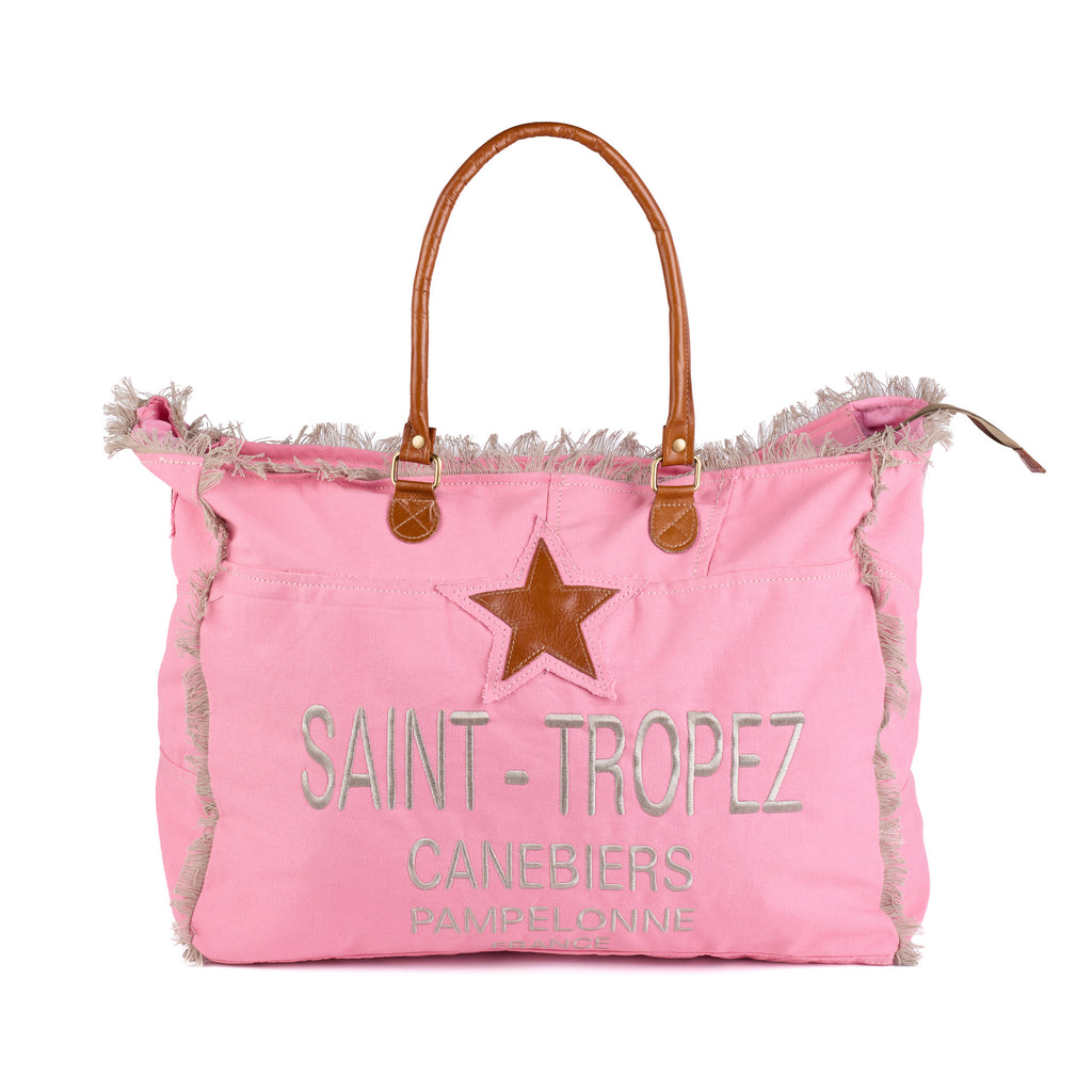 Casey: a pink washed canvas beach bag with frayed raw edges, a tan leather star patch and embroidered Saint-Tropez Canebiers Pampelonne France lettering, front view with the handles raised