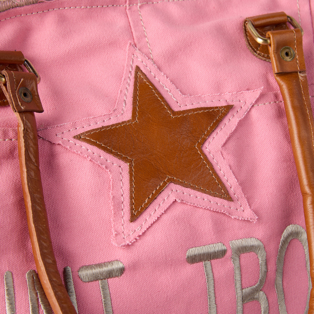 Casey: a pink washed canvas beach bag with frayed raw edges, a tan leather star patch and embroidered Saint-Tropez Canebiers Pampelonne France lettering, shown at an angle
