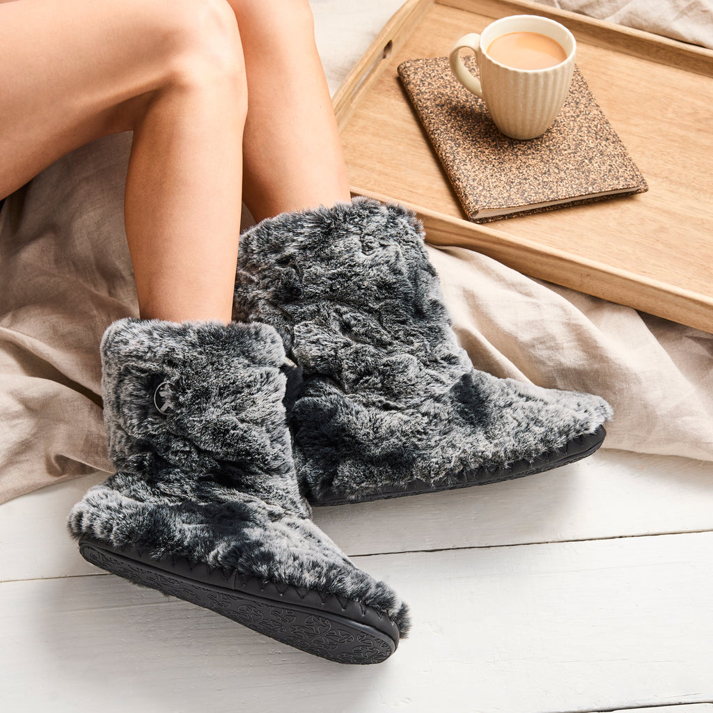 A person wearing plush gray slipper boots sits on a white wooden floor beside a wooden tray holding a mug of coffee.