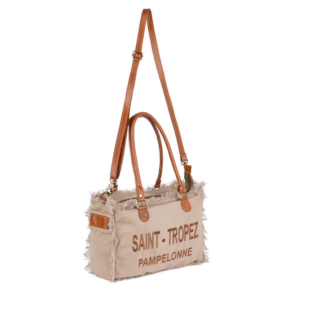 Lani: a beige washed canvas shoulder bag with frayed raw edges, tan leather trim and embroidered Saint-Tropez Pampelonne lettering, shown in full from an angle