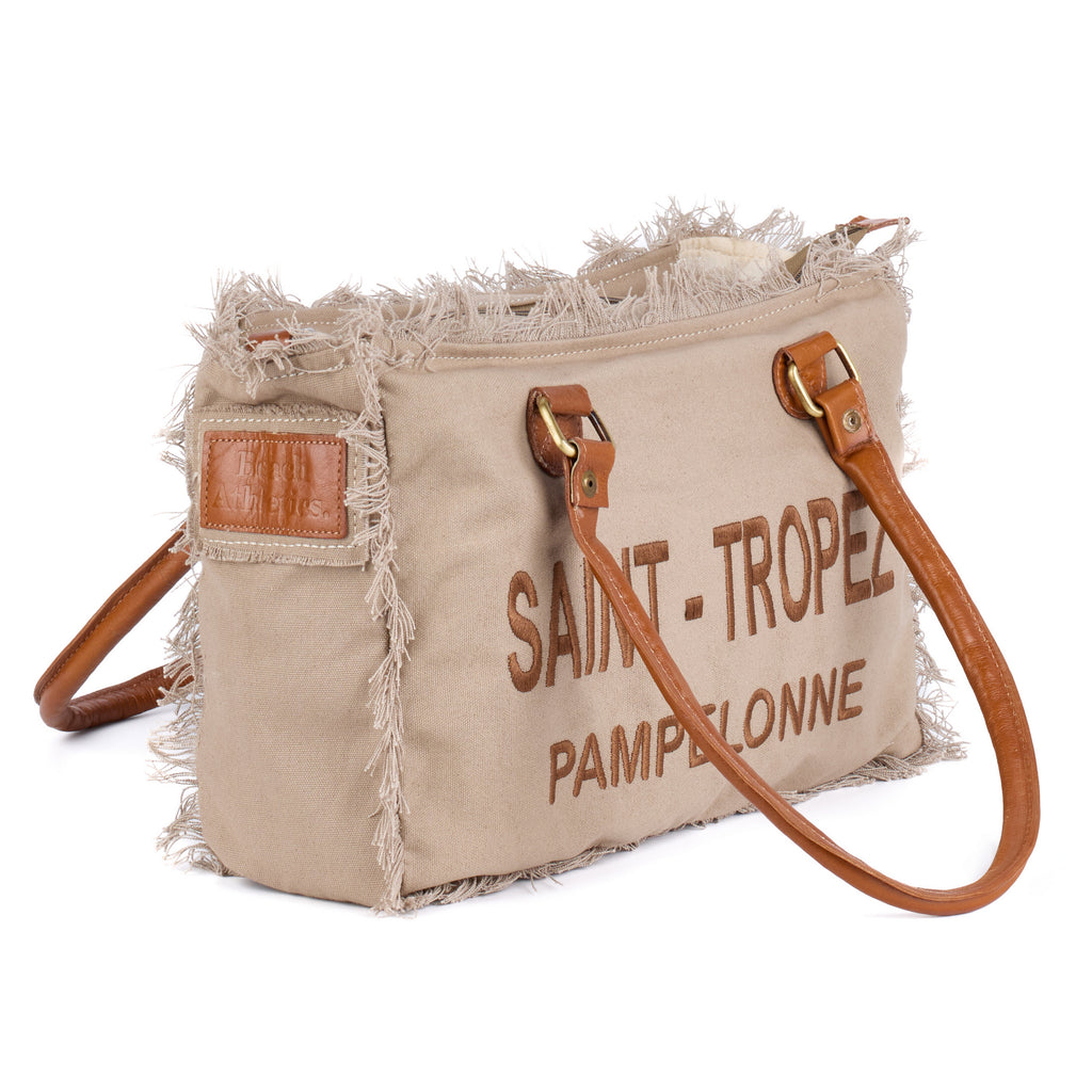 Lani: a beige washed canvas shoulder bag with frayed raw edges, tan leather trim and embroidered Saint-Tropez Pampelonne lettering, shown at an angle