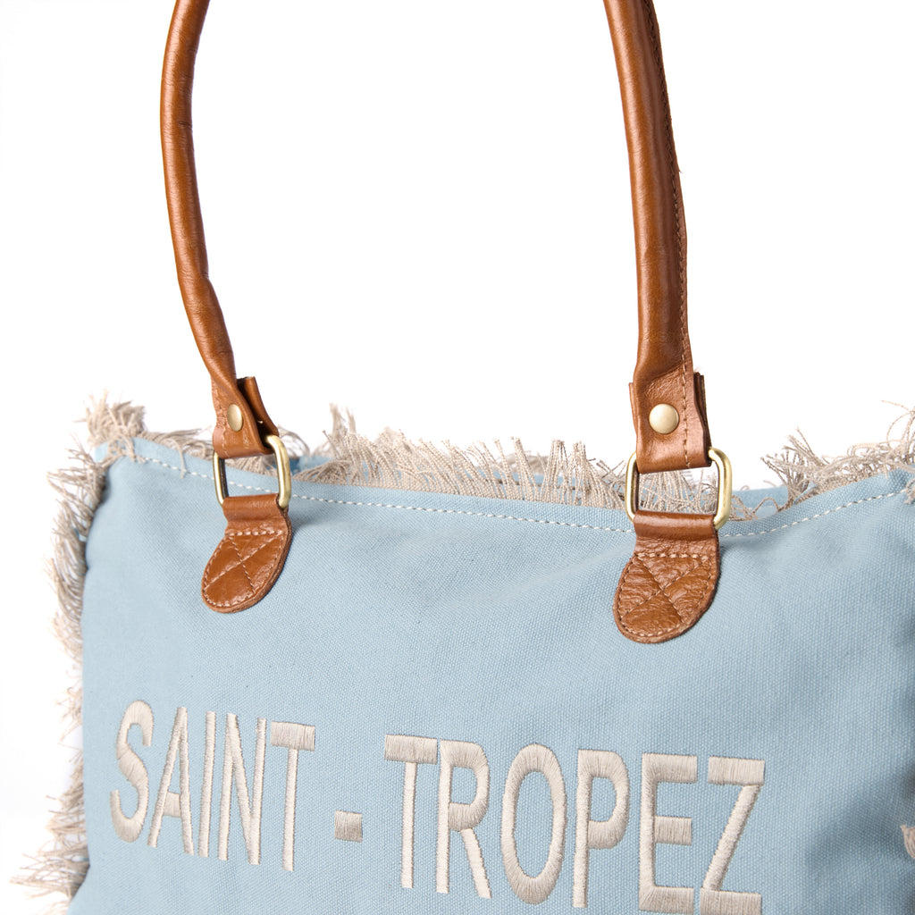 Lani: a blue washed canvas shoulder bag with frayed raw edges, tan leather trim and embroidered Saint-Tropez Pampelonne lettering, shown at an angle