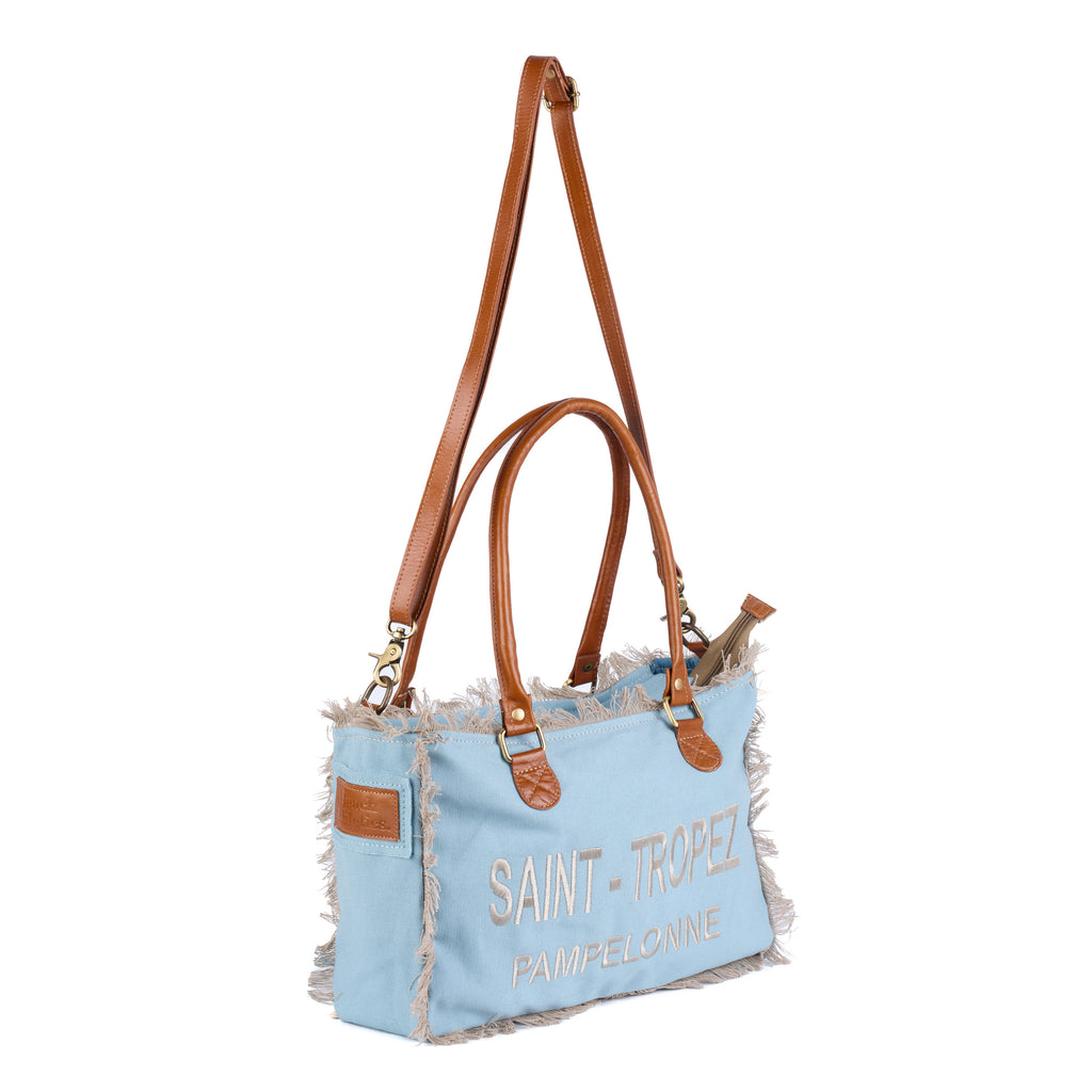 Lani: a blue washed canvas shoulder bag with frayed raw edges, tan leather trim and embroidered Saint-Tropez Pampelonne lettering, shown in full from an angle