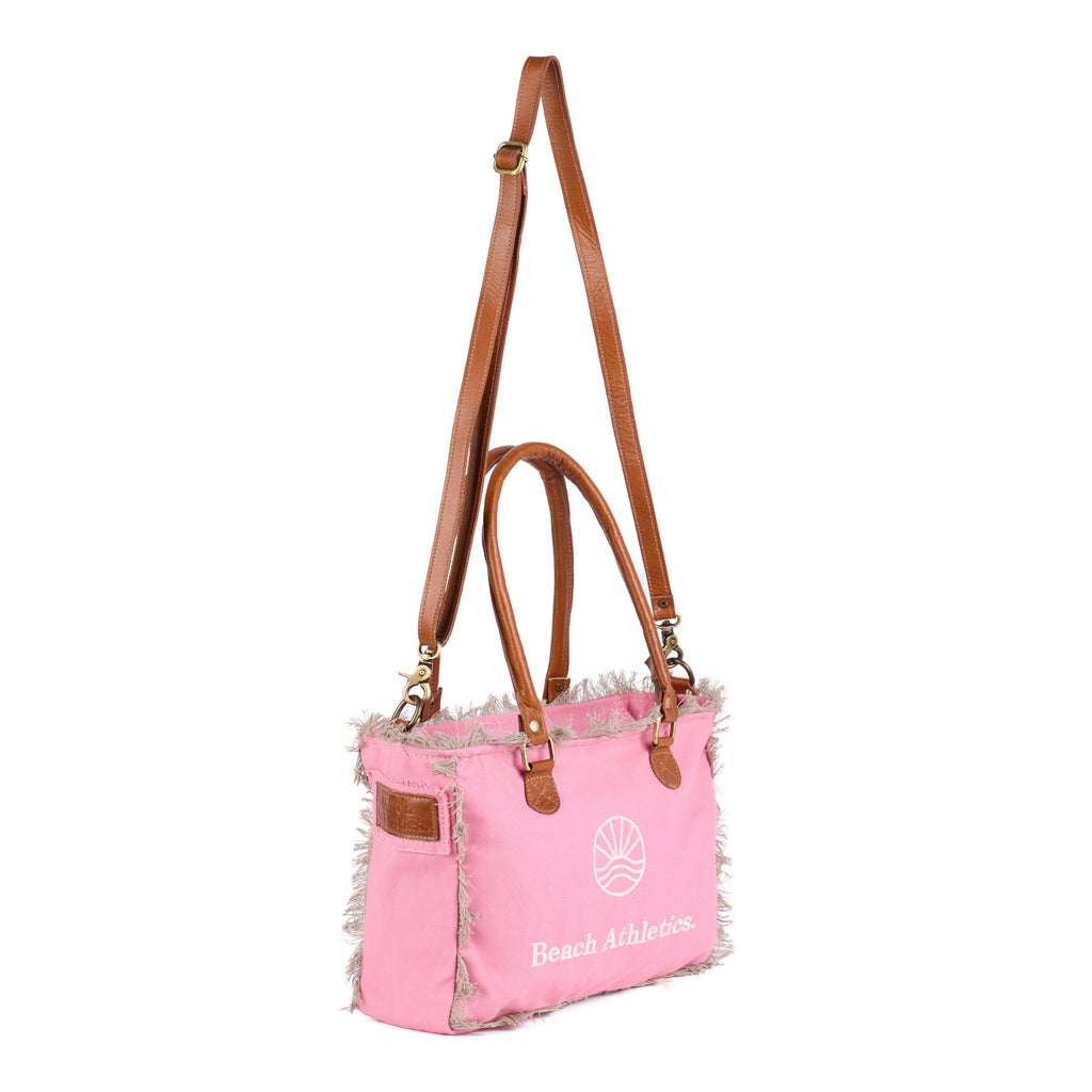 Shauni: a pink washed canvas shoulder bag with frayed raw edges, tan leather trim and an embroidered sunrise Beach Athletics logo, shown in full from an angle